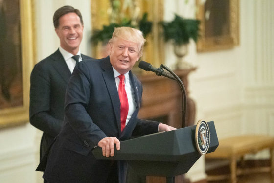 Mark Rutte laughing in the background as Donald Trump leans over to the side of the White House podium.